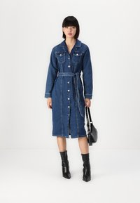 Τζιν φόρεμα σε σκούρο μπλε, διαθέτει μακριά μανίκια, κουμπιά μπροστά, διπλές τσέπες στο στήθος και ζώ�νη στη μέση που δένει. Συνδυάζεται με μαύρες μπότες.