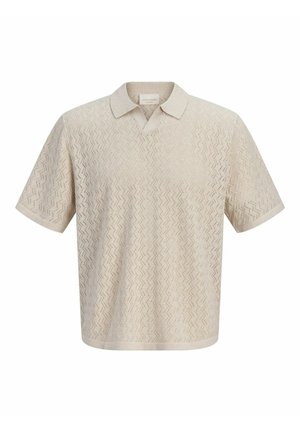 Mosolygó fiú áll, bézs texturált rövid ujjú pólót, kék farmer sortot, fehér zoknit és barna bőr cipőt visel egyszínű háttér előtt.