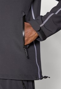 Jaqueta preta com um bolso com fecho éclair, fecho de pulso em velcro e costura branca em contraste. Textura suave com bainha ajustável.
