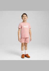 Rožinė medvilninė marškinėliai su balta „PUMA“ logotipu ir derančiomis šortais, kartu su oranžiniais batais ir baltais kojiniais, pateikti neutralioje fone.