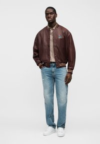 Άνδρας ντυμένος με καφέ δερμάτινο μπουφάν τύπου bomber, μπεζ πόλο πουκάμισο, ανοιχτό μπλε τζιν και λευκά αθλητικά παπούτσια, στέκεται μπροστά σε λευκό φόντο.