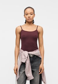 Νεαρή γυναίκα με πλεξούδες φοράει μπορντό αμάνικο τοπ, γκρίζα τζιν με ζώνη που έχει αγκράφα σε σχήμα καρδιάς και μια ριγέ μπλούζα δεμένη γύρω από τη μέση της.