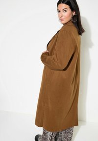 Bruine oversized jas van zachte stof, met een geknipt kraag, zijzakken en een gladde textuur; gedragen over een patroonbroek.