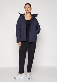 Granatowa kurtka z kapturem na zamek z przodu, dużymi kieszeniami i elastycznymi mankietami, zestawiona z czarnymi spodniami z zamkami oraz białymi trampkami.