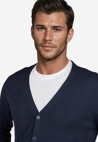 Dunkelblauer Cardigan mit V-Ausschnitt, gerippter Textur und drei blauen Knöpfen. Kombiniert mit einem weißen Rundhals-T-Shirt.