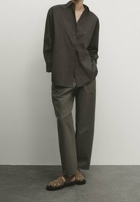 Dunkelblaues Hemd mit langen Ärmeln, kombiniert mit lockeren, olivgrünen Hosen und braunen Sandalen mit Riemen. Neutraler Hintergrund und Boden.