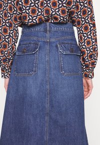 Personne portant une jupe en jean bleue avec des poches à rabat boutonnées à l’arrière et une chemise à manches longues à motifs rentrée à l’intérieur.
