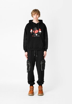 Joven con cabello corto rubio, vistiendo una sudadera negra con un gráfico rojo y blanco, pantalones de chándal cargo negros y botas de cordones color beige, de pie.