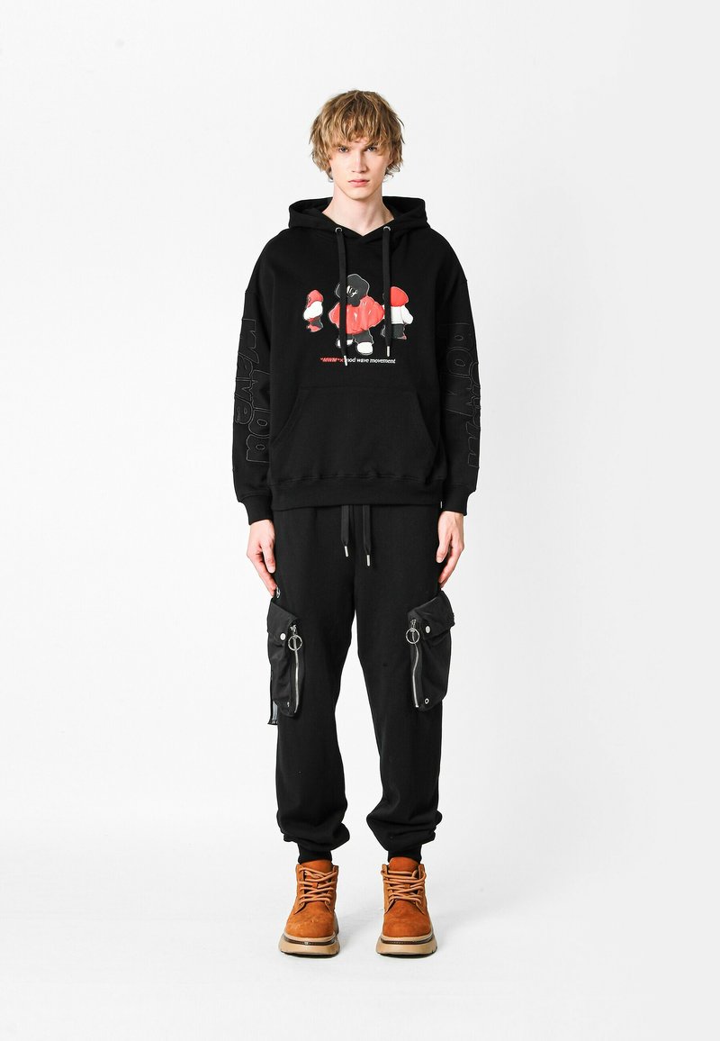 Joven con cabello corto rubio, vistiendo una sudadera negra con un gráfico rojo y blanco, pantalones de chándal cargo negros y botas de cordones color beige, de pie.