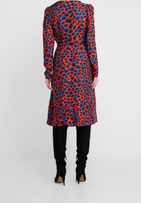 Långärmad klänning i ett livligt leopardmönster i röd, blå och svart. Har en cinchad midja och en ventil bak. Bärs över svarta trånga byxor och stövlar.