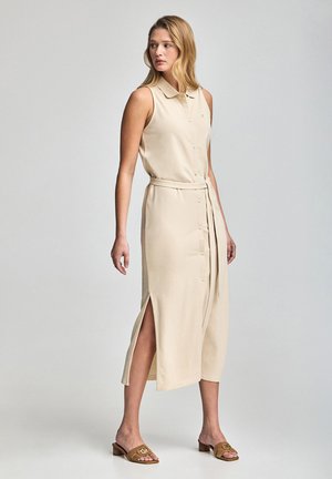 Abito beige senza maniche con colletto, bottoni sul davanti, fessure laterali e vita legata. Il materiale sembra liscio e testurizzato.