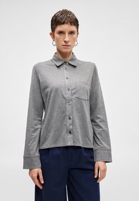 Γκρίζο πουκάμι�σο με μακριά μανίκια και γιακά, κουμπώνει μπροστά, έχει μία τσέπη στο στήθος και φαρδιά μανσέτα. Μαλακό ύφασμα με λεία υφή.