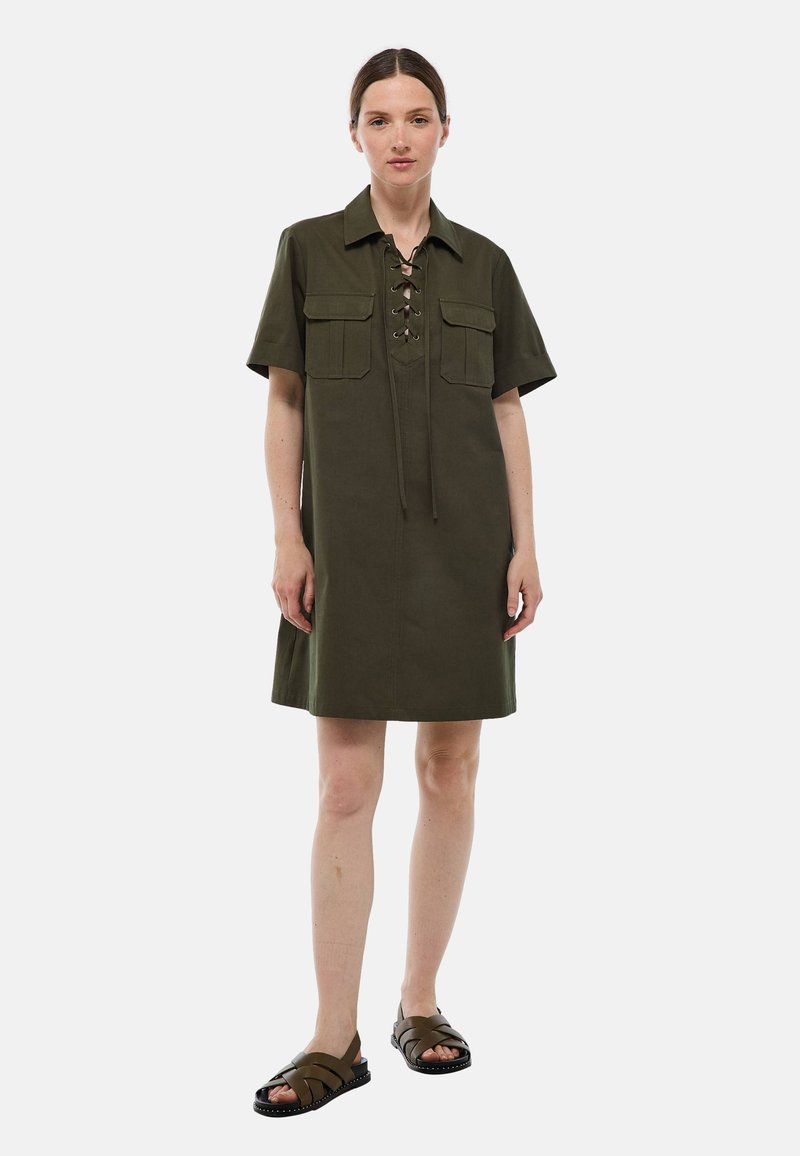Robe à manches courtes kaki avec un décolleté à lacets et deux poches poitrine. Le tissu semble lisse, et le design est décontracté et droit.