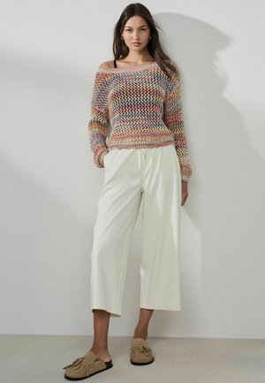 Woman wearing multicolored knit sweater, white wide-leg pants, and beige tasseled slip-on shoes standing against a plain light wall.