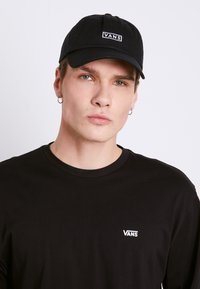 Cappellino Vans nero con un design strutturato e logo bianco, indossato con una maglietta a maniche lunghe nera con un piccolo logo sul petto.