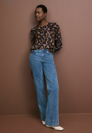 Hnedo-čierny a béžový top s leopardím vzorom na dlhé rukávy, spárovaný so svetlo modrými džínsy s širokými nohavicami. Model má na sebe krémové topánky na špičke.