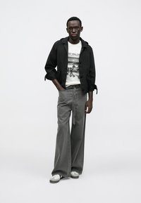 Čierna bunda cez biele tričko s potlačou, sivé nohavice s širokými nohavicami na sťahovanie a biele tenisky so sivými detailmi. Minimalistický dizajn.