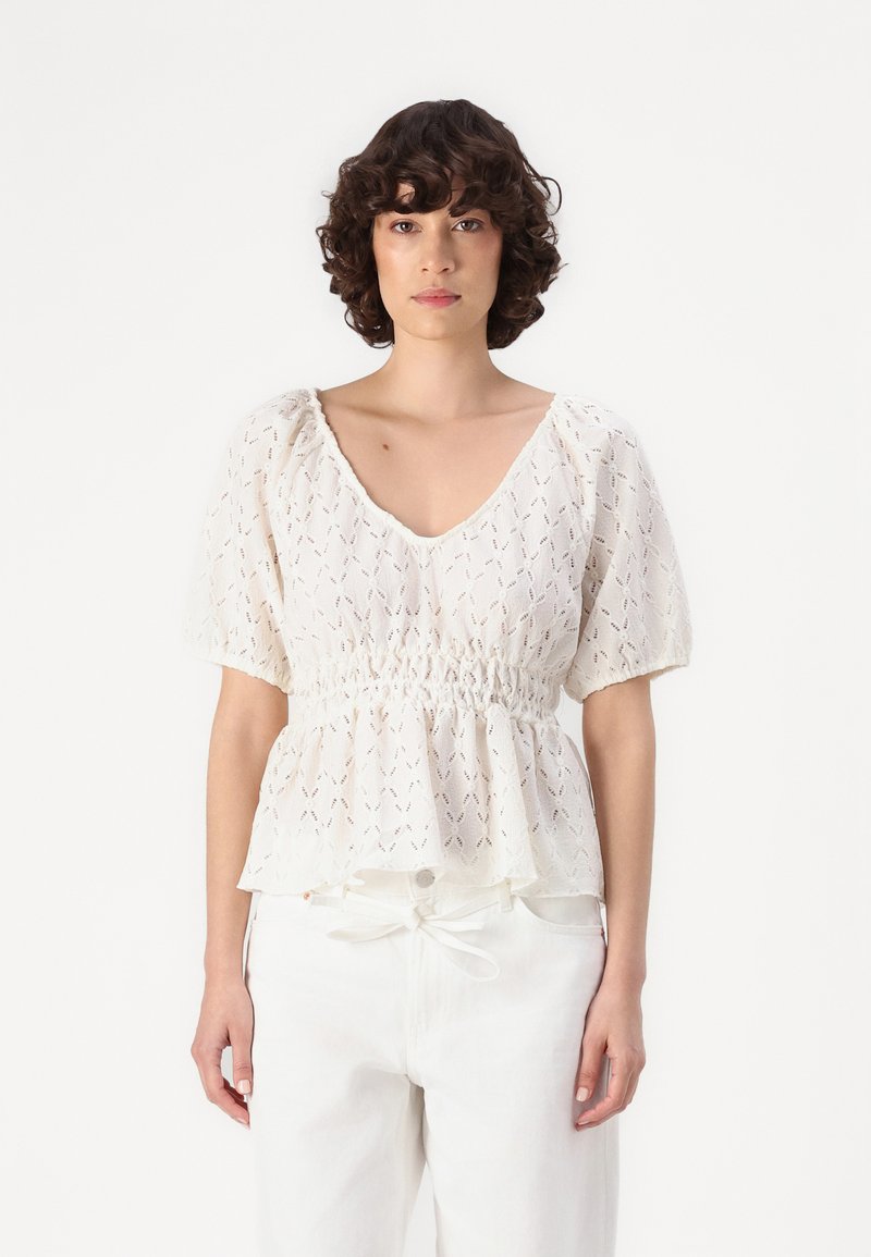Chemisier blanc à manches courtes avec un col en V, taille smockée et motif en dentelle. Associé à un pantalon blanc à cordon de serrage.
