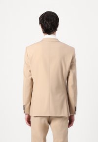 Veste de costume beige avec des épaules structurées, des revers crantés et deux boutons. Tissu lisse, coupe ajustée et fentes arrière doubles pour le mouvement.