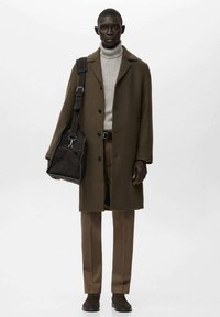 Abrigo de lana marrón sobre un suéter gris de cuello alto, combinado con pantalones marrones y zapatos oscuros. Un bolso tipo mensajero oscuro está colgado al hombro.