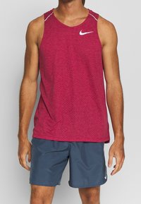 Rotes Sporttanktop mit kleinen Perforationen, weißem Nike-Logo, kombiniert mit grauen Shorts aus feuchtigkeitsableitendem Material.