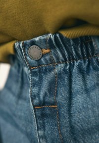 Jeans mają marszczony pas, kontrastowe pomarańczowe szwy oraz okrągły czarny metalowy guzik z wyrytym napisem "Nineteen Eighty".