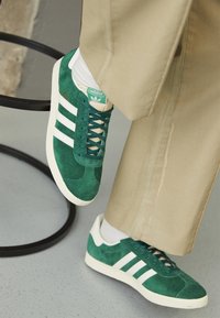 Green suede sneakers with white stripes, featuring a textured surface, rounded toe, and branded tongue. Paired with beige pants.