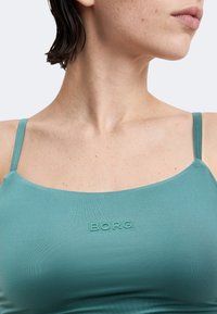 Γυναίκα που φοράει τοπ τιρκουάζ με λεπτές τιράντες, που δείχνει τον λαιμό, τους ώμους και το πάνω μέρος του στήθους, με το λογότυπο "BORG" μπροστά.
