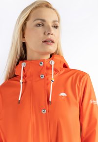 Oranje waterdichte jas met een capuchon, voorzien van witte trekkoorden, drukknoopsluitingen en een klein paraplu-logo op de borst. Gladde textuur.
