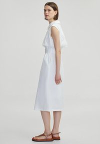 Witte jurk met een getailleerde taille, mouwloos ontwerp en geborduurd schouderdetail. Draagt met bruine sandalen. Glad, lichtgewicht stof.