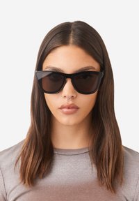 Black oversized sunglasses with dark lenses and angular frames, worn by a person with shoulder-length dark hair, against a white background.