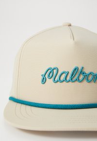 Beige caps med en sydd turkise logo "Malbon." Har et teksturert bånd og en buet skjerm. Glatt stoff med synlige sømmer.