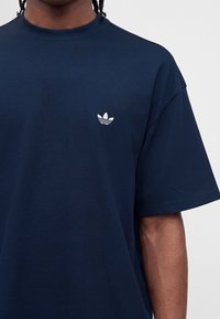 Σκούρο μπλε μπλούζα με το λευκό λογότυπο Adidas trefoil στο αριστερό στήθος, φορεμένη από άτομο με σκούρο δέρμα και πλεξούδες.