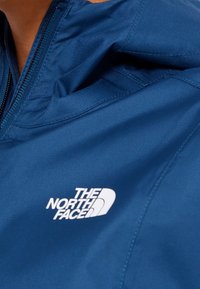 Niebieska kurtka z materiału z zamkiem błyskawicznym z przodu, z białym logo "The North Face" na lewej stronie klatki piersiowej. Gładka tekstura, casualowy styl.