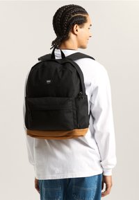 Svart tygsryggsäck med brun botten, ett enda framficka och axelremmar. Har en logotypmärke på framsidan.