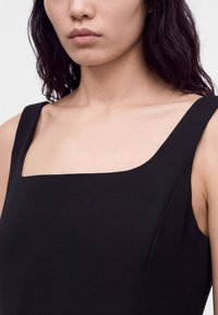 Femme aux cheveux foncés mi-longs portant un haut noir sans manches à encolure carrée sur un fond clair uni.