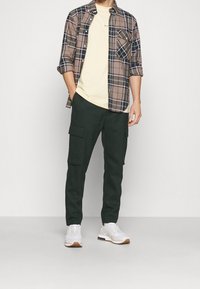 Rento asukokonaisuus, jossa ruskea ruutupaita vaaleankeltaisen t-paidan päällä, yhdistetty tummanvihreisiin cargo-housuihin ja valkoisiin urheilukenkiin.