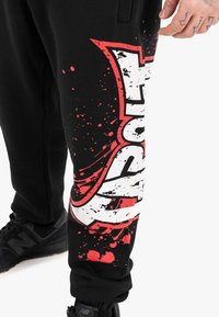 Schwarze Jogginghose mit einem großen weißen und roten Grafikdesign sowie Spritzmustern. Mit elastischen Bündchen und seitlichen Taschen.