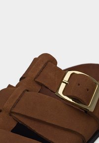 Bride de sandale en daim marron avec une boucle rectangulaire dorée, montrant les coutures et les trous de réglage sur une semelle plate.