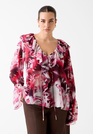 Kobieta o ciemnych włosach w różowej, czerwonej i białej kwiecistej bluzce z falbanami oraz brązowych spodniach, stojąca na tle jednolitego białego tła.