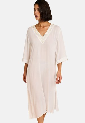 Femme portant une robe ample, longueur genou, de couleur crème, manches 3/4 et col en V orné, debout devant un fond clair uni.