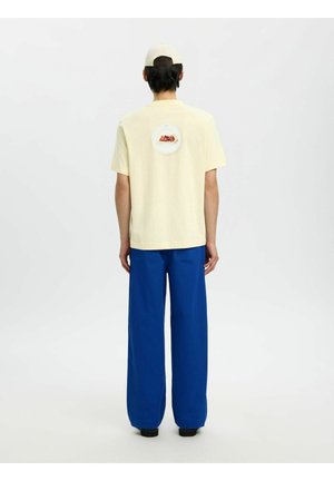 Mężczyzna ubrany w kremową koszulkę z okrągłym nadrukiem na plecach, niebieskie szerokie spodnie, czarne buty i kremową czapkę, stojący na białym tle.