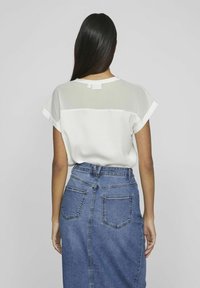 Kvinna med långt mörkt hår som bär en vit blus med korta ärmar och en höga midjan blå denimkjol, sedd bakifrån mot en enkel bakgrund.