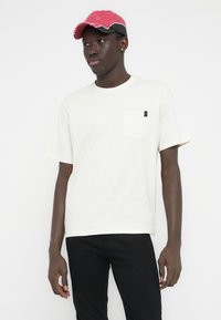 Camiseta blanca de manga corta con un bolsillo en el pecho y una pequeña etiqueta marrón. Combinada con jeans negros y una gorra rosa con detalles en negro.