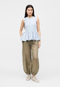Kvinne iført lyseblå ermeløs peplum-topp med glidelås foran, olivengrønne vide bukser med rynkede mansjetter og beige joggesko, stående mot hvit bakgrunn.