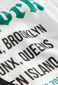 Katoenen T-shirt met gedurfde zwarte en groene grafische tekst. De achtergrond is zacht wit met verschillende lettergroottes en stijlen, inclusief plaatsnamen.