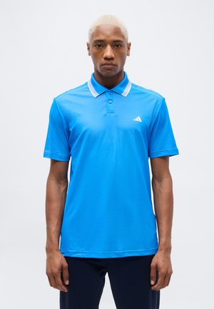 Άνδρας με κοντά ξανθά μαλλιά που φοράει μπλε polo μπλούζα Adidas με λευκές λεπτομέρειες στον γιακά, στέκεται μπροστά σε λευκό, απλό φόντο.