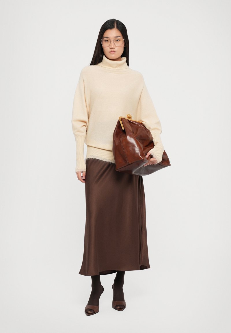 Pull en col roulé beige, jupe en satin marron, sac en cuir marron avec des accessoires dorés, et collants marron avec des escarpins ouverts.