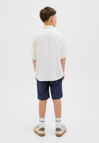 Bílé tričko s krátkým rukávem a zadním pasem, doplněné tmavě modrými šortkami. Tento outfit zahrnuje béžové tenisky a bílé ponožky s černým potiskem.