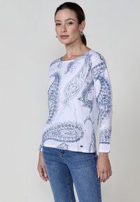 Mujer de pie que lleva una camisa blanca de manga larga con patrones de paisley azules y jeans azules, contra un fondo claro y sencillo.